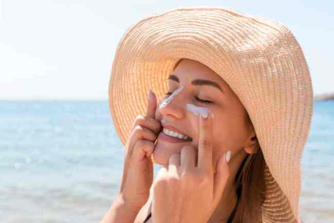 Smiling woman in a hat at beach applying sunblock to face Smiling woman in a hat at beach applying sunblock to face