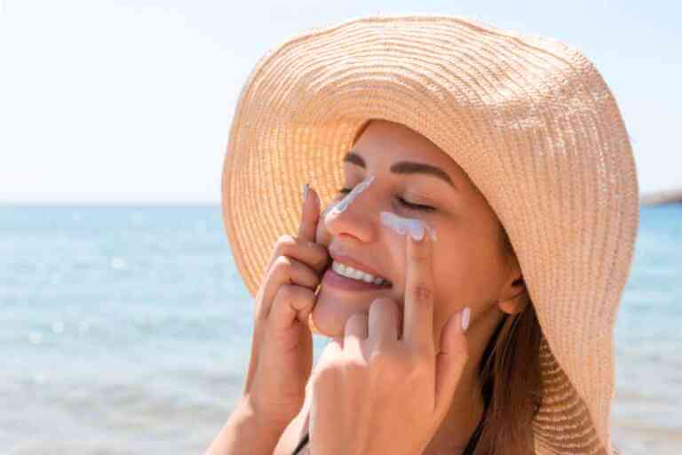 Smiling woman in a hat at beach applying sunblock to face